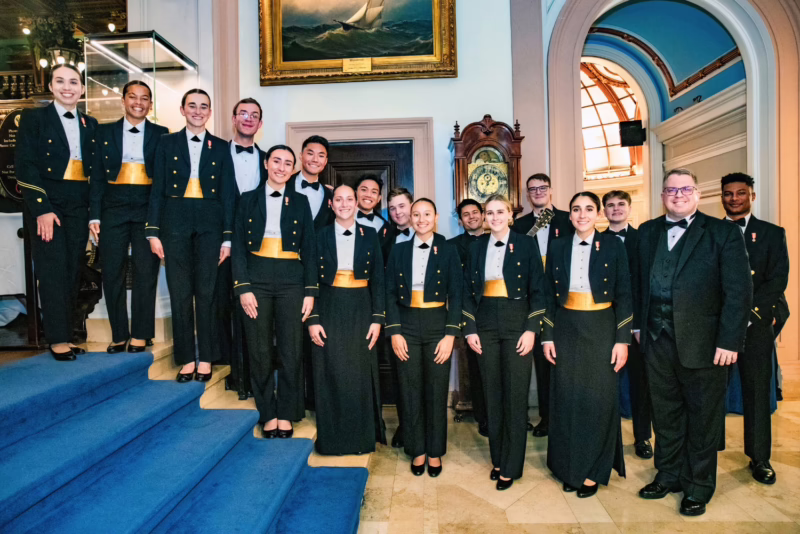 Group photo of the Coast Guard Cadet Chorale in formal attire, standing together on a blue-carpeted staircase in an elegant, historic setting. The cadets are dressed in dark uniforms with gold sashes, and they are accompanied by their director, posing in front of a beautifully detailed clock and maritime-themed artwork.
